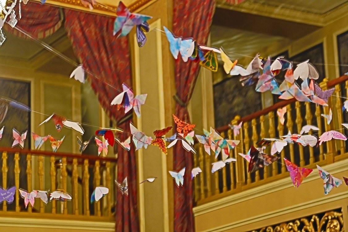 Paper birds hang above an ornate theater balcony and red curtains.