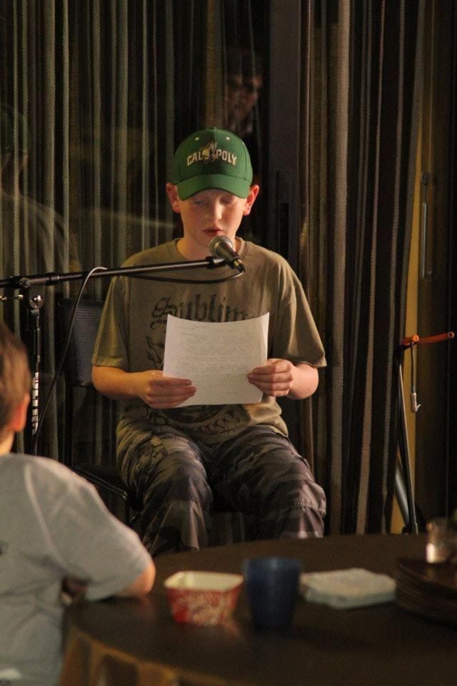 A seated adult reads from a sheet of paper onstage.