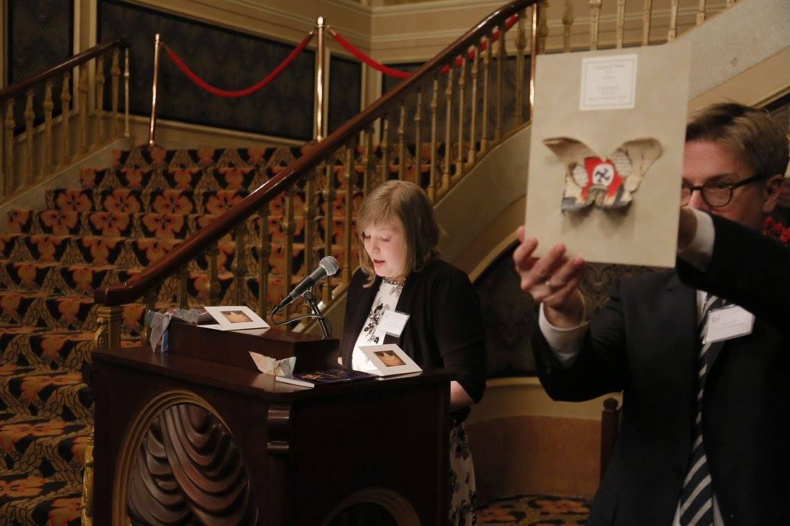 An adult reads at a lectern while someone holds up a small artwork.