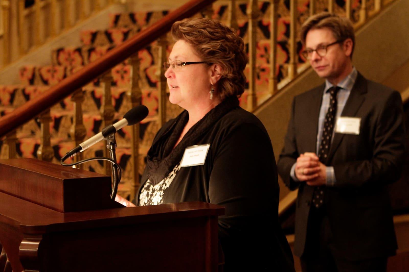 An adult speaks at a lectern while an adult stands nearby.