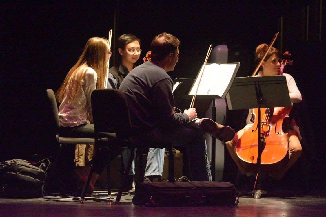 Musicians perform with piano and string instruments on a dark stage.