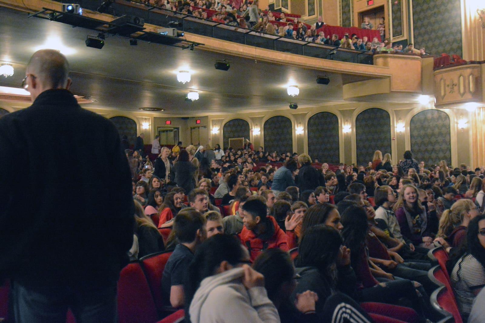 A speaker faces a large seated theater audience.