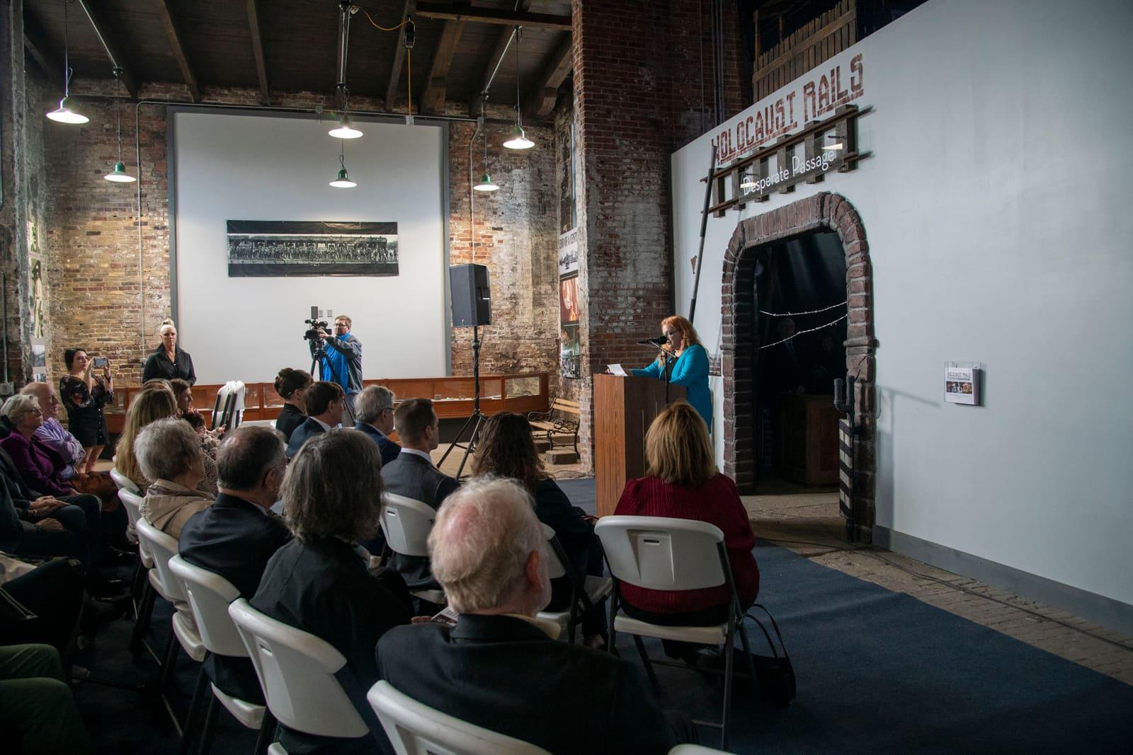 An audience listens in a brick-walled hall.