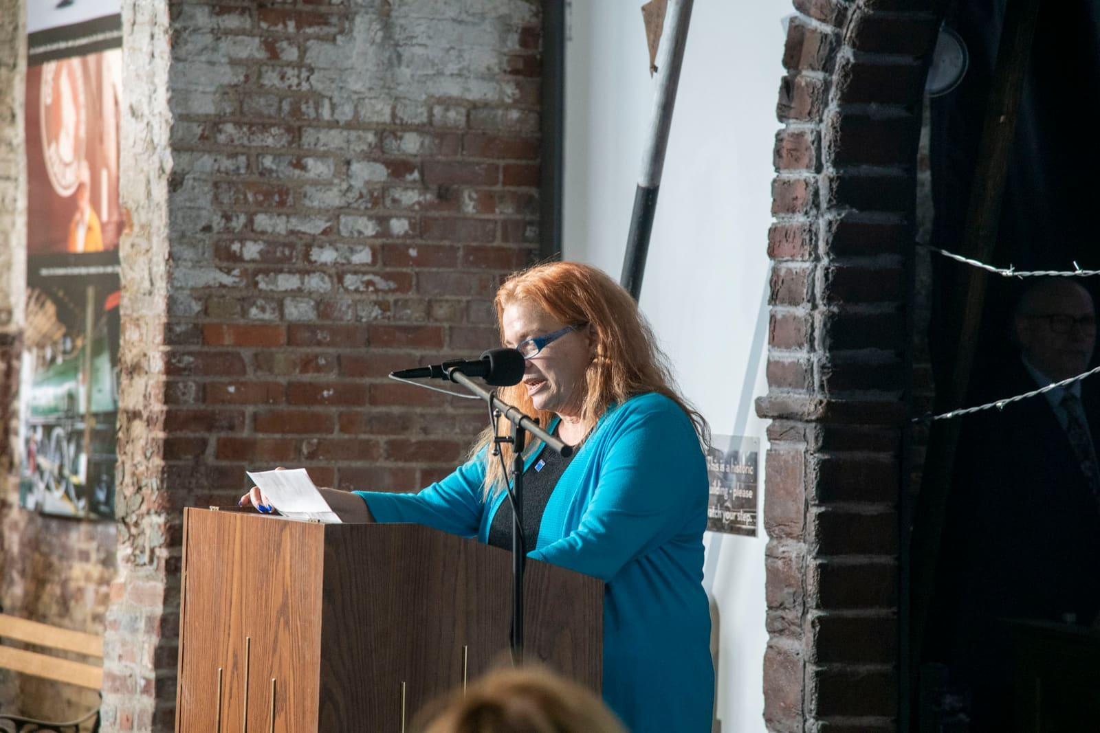 An adult reads from a wooden podium in a brick-walled venue.