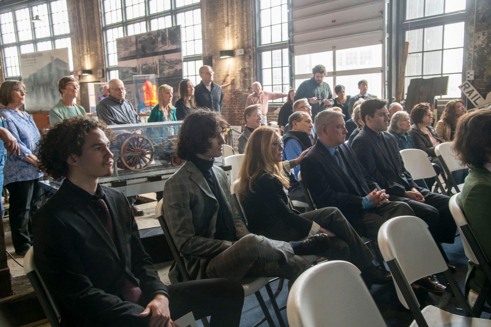 An audience sits on folding chairs in a sunlit industrial room.