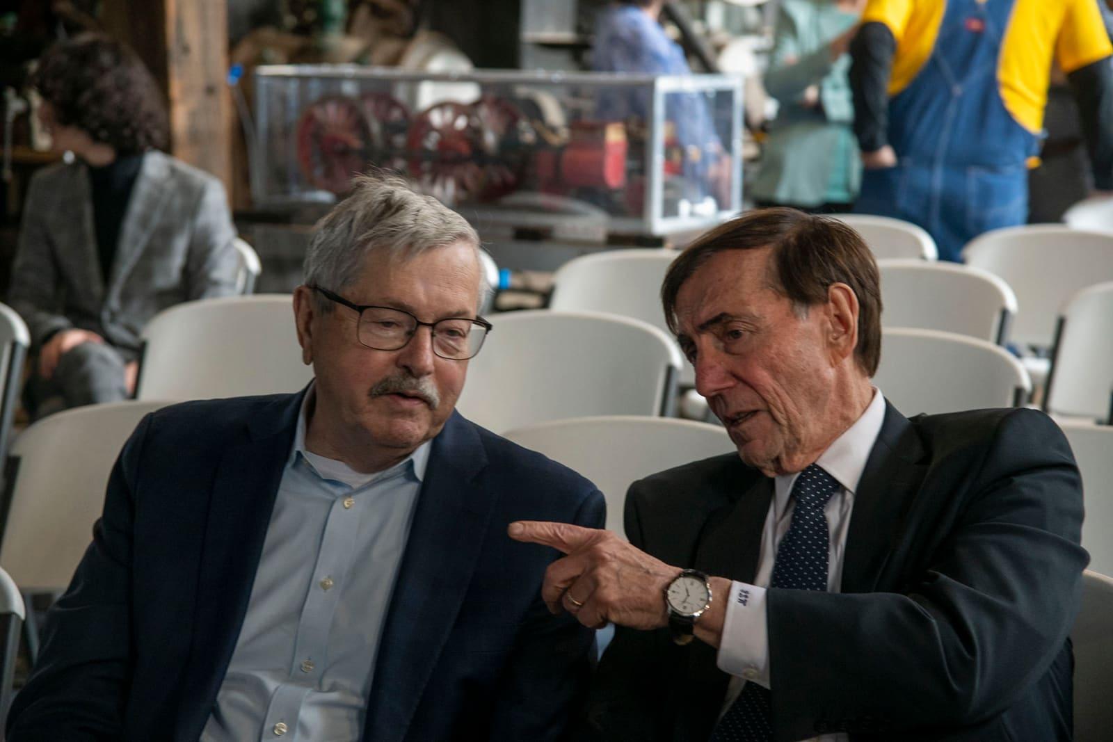 Two adults converse while seated among rows of white chairs.