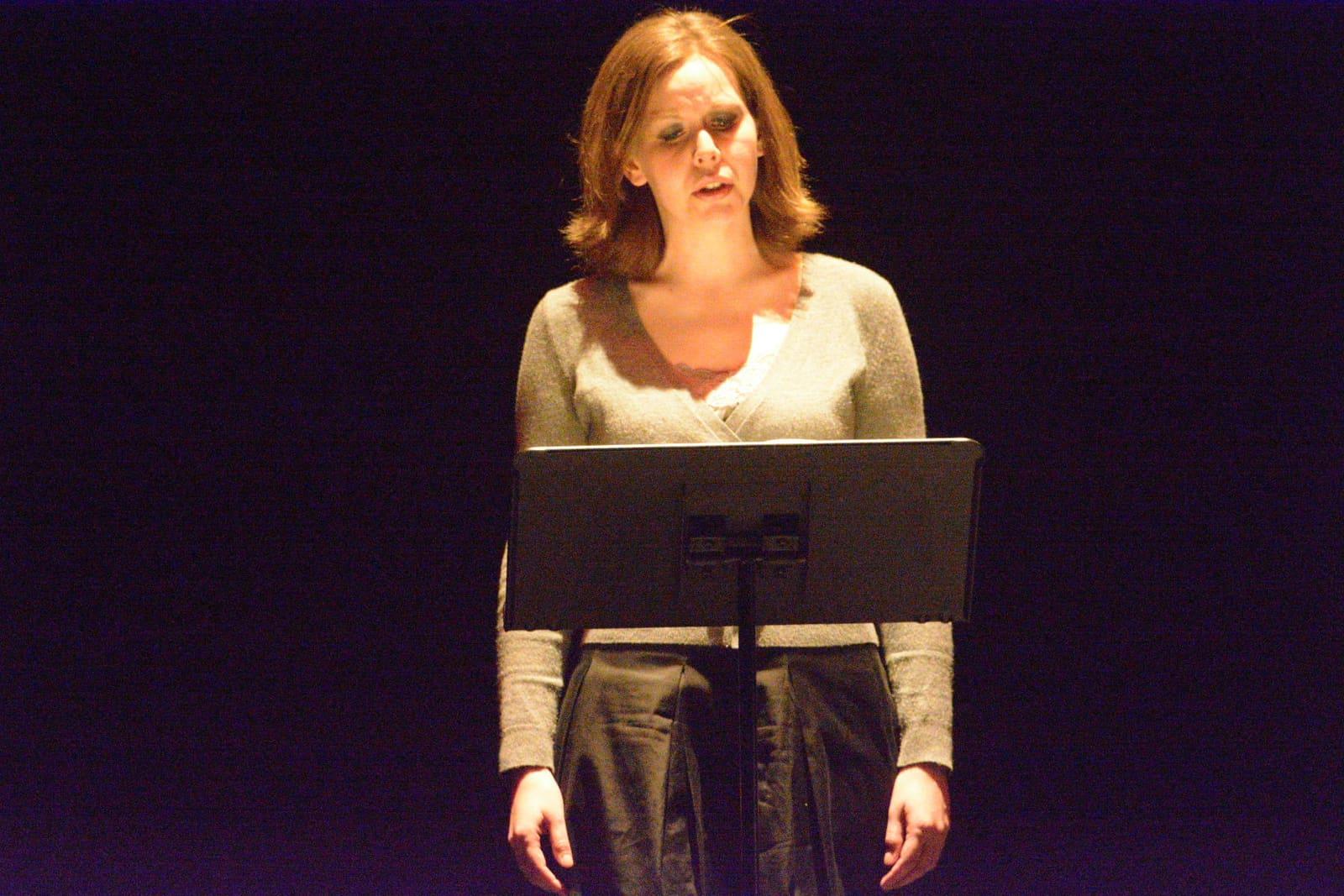 An adult sings from behind a music stand onstage.