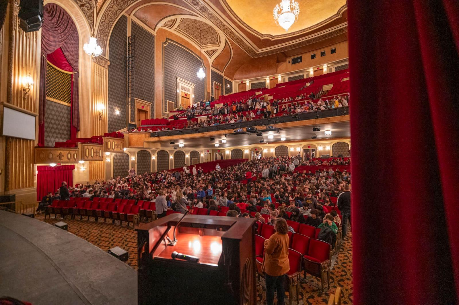 A packed theater is viewed from behind the stage curtain.