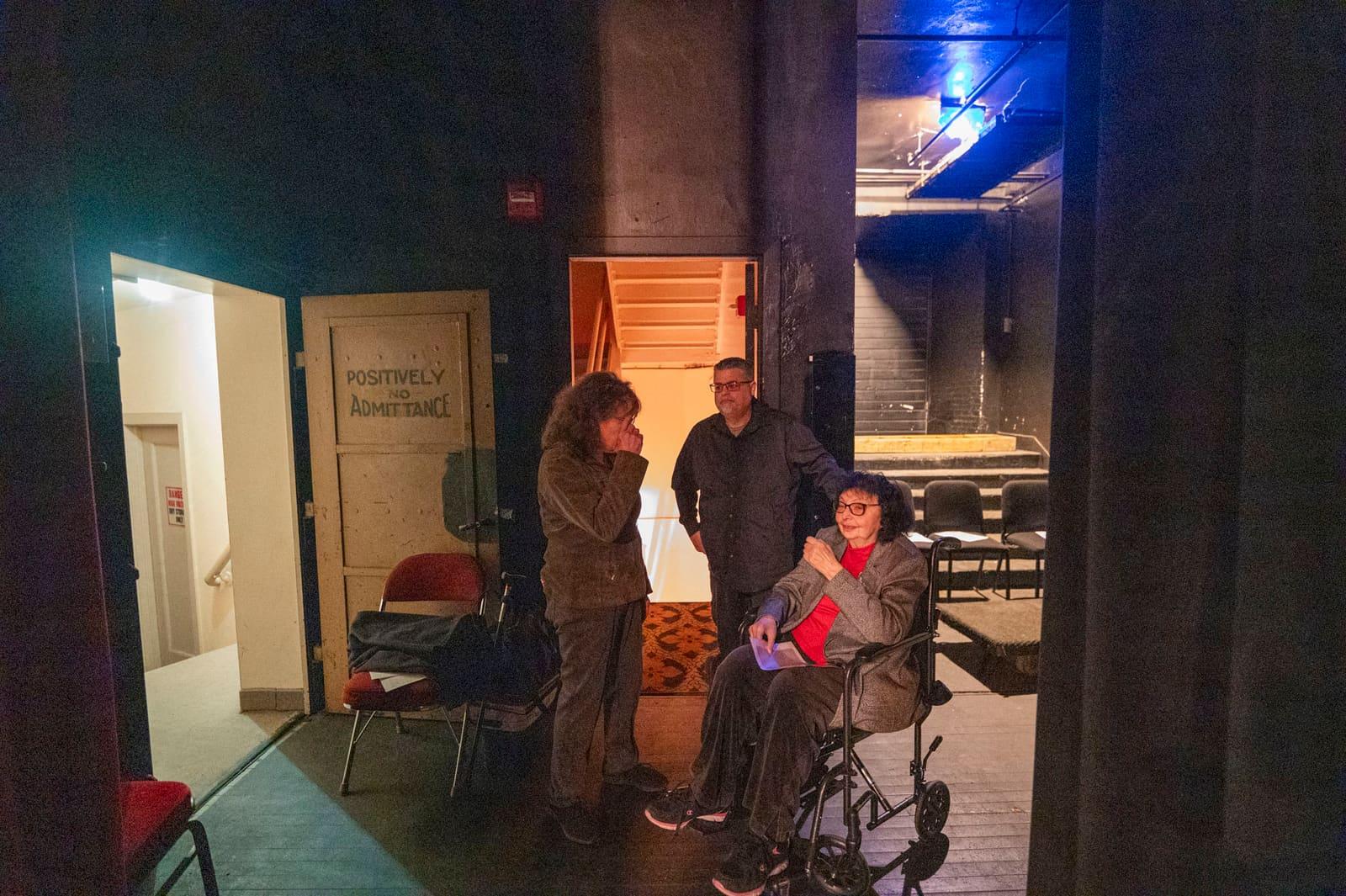 Three adults gather backstage, including one seated in a wheelchair.
