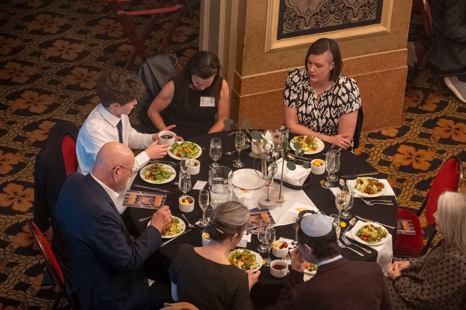 Six adults sit around a round table with plated meals.