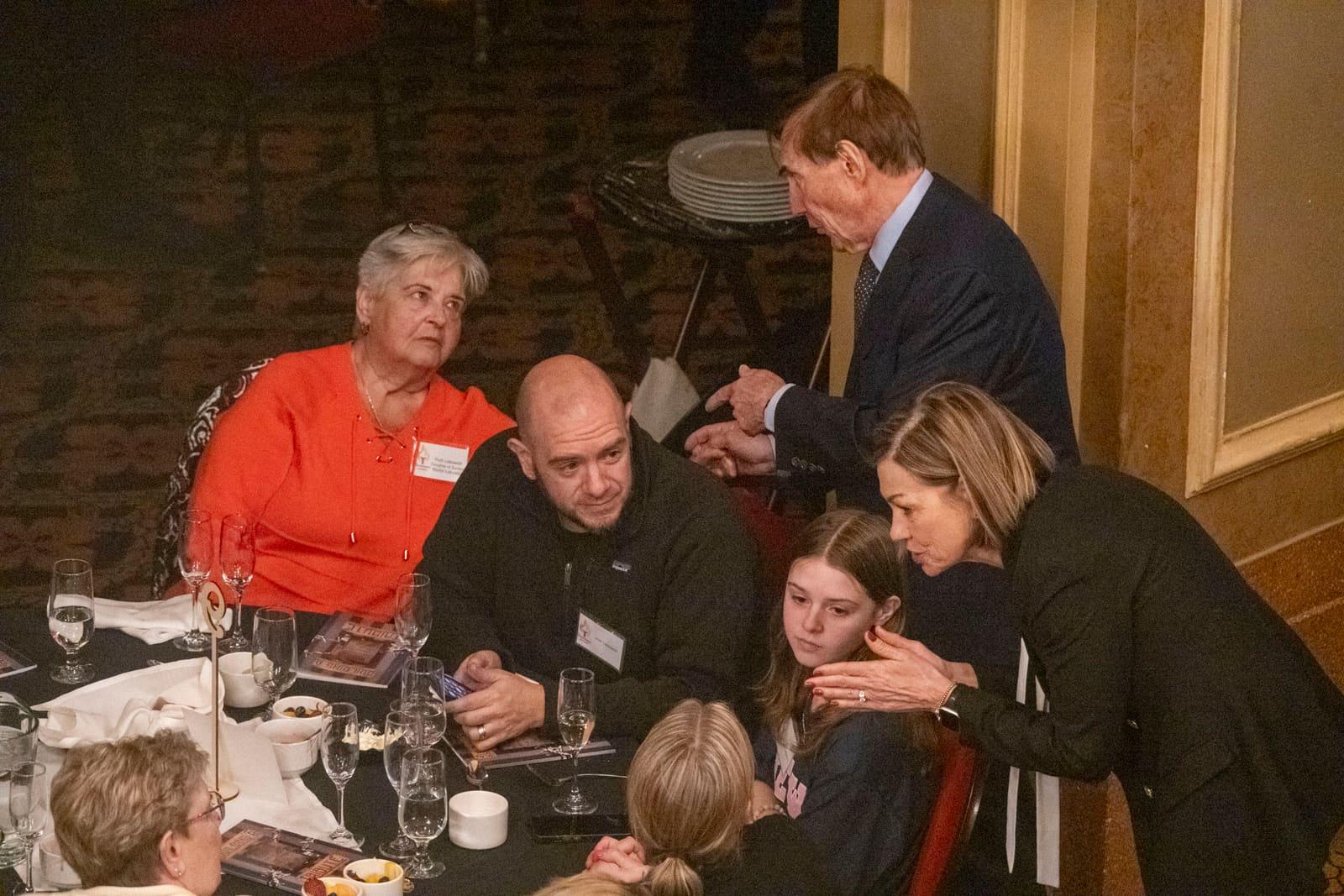 Several people lean toward a seated guest at a banquet table.