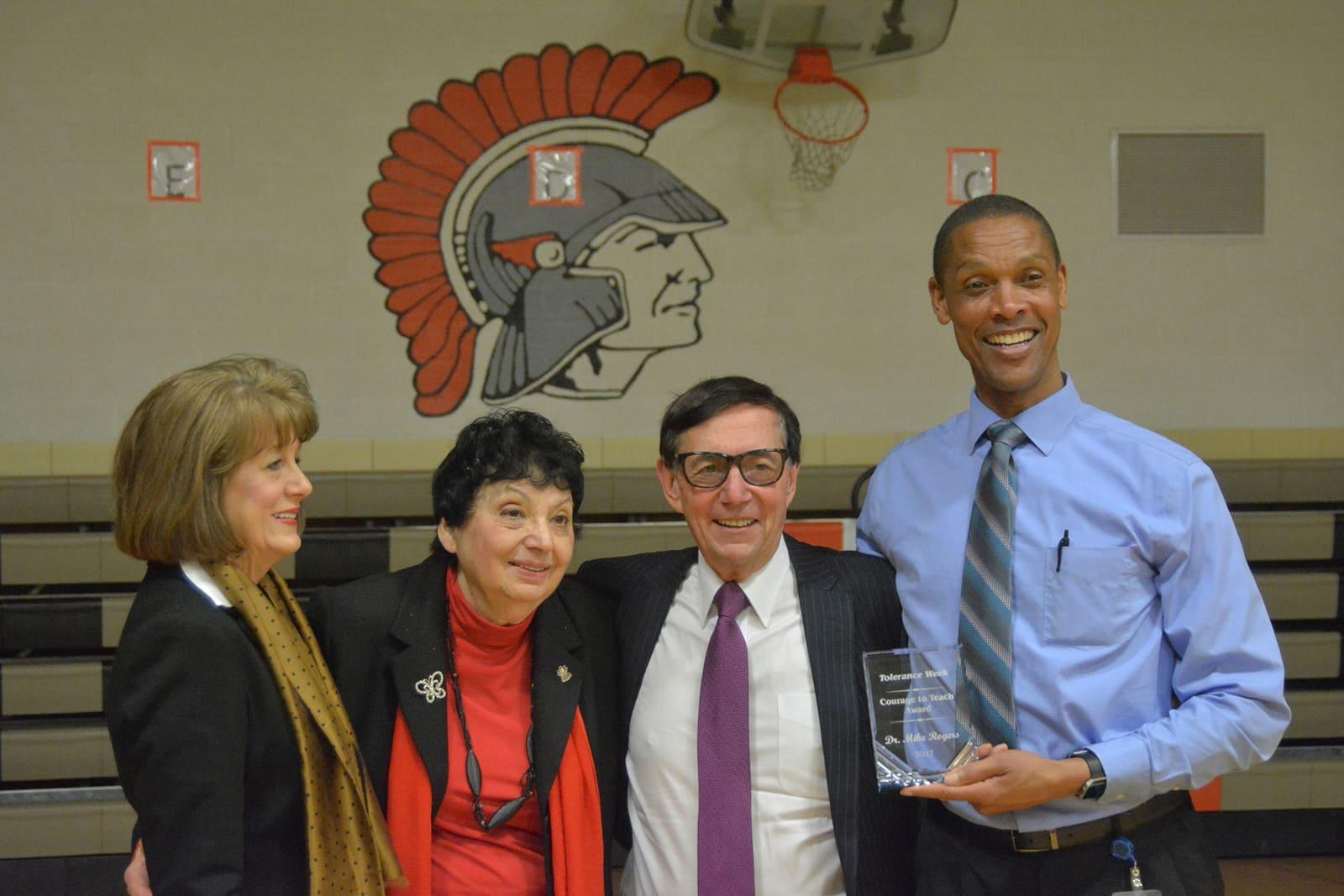 Four adults pose in a gymnasium while one holds a clear rectangular award.