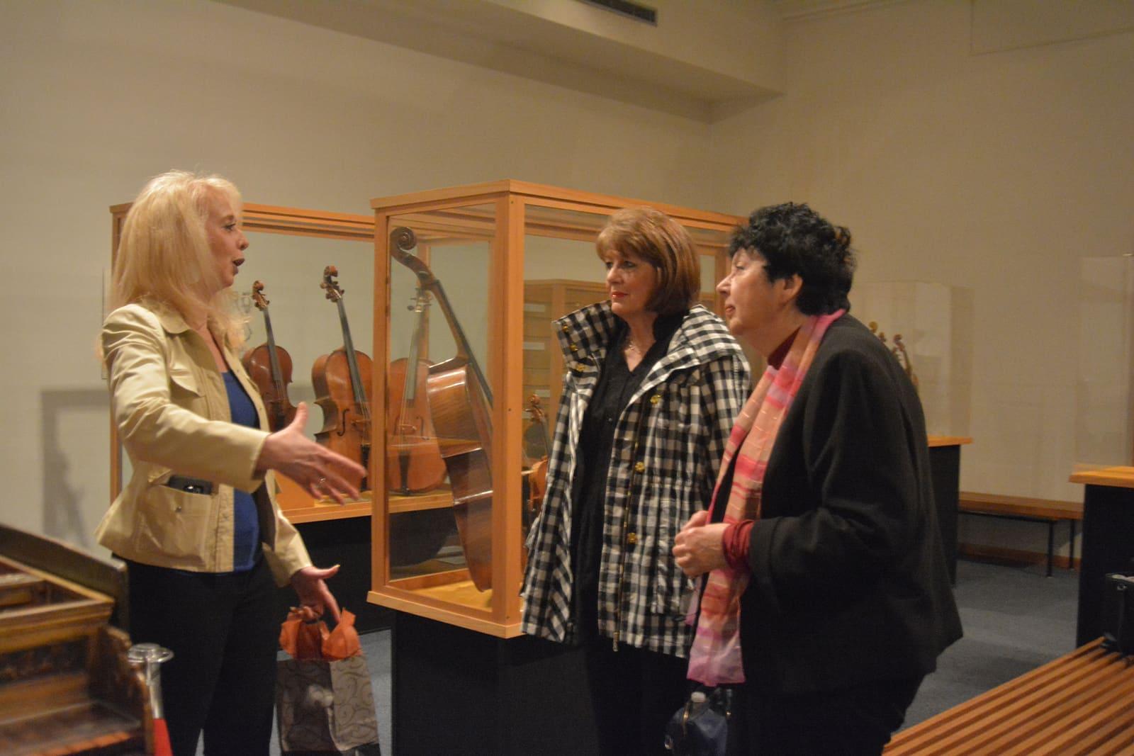 Three women talk beside a glass instrument case in a museum gallery.