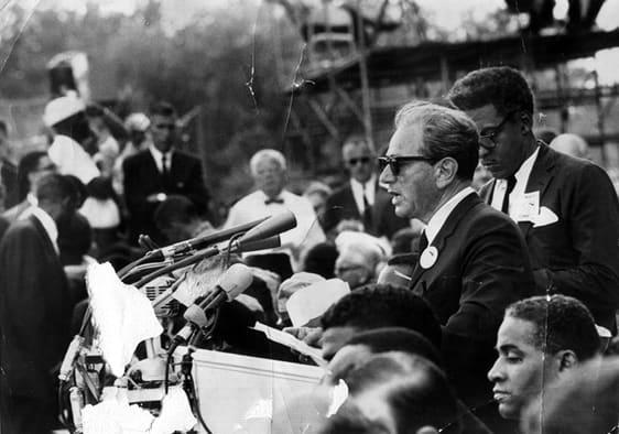 Black-and-white photograph of a man speaking into several outdoor microphones before a seated crowd.