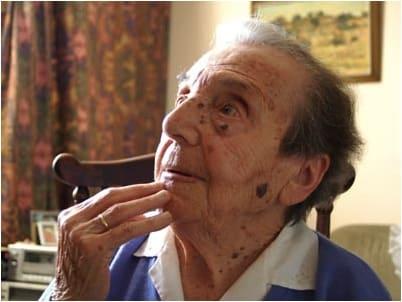 An older woman sits in profile with one hand raised near her mouth.