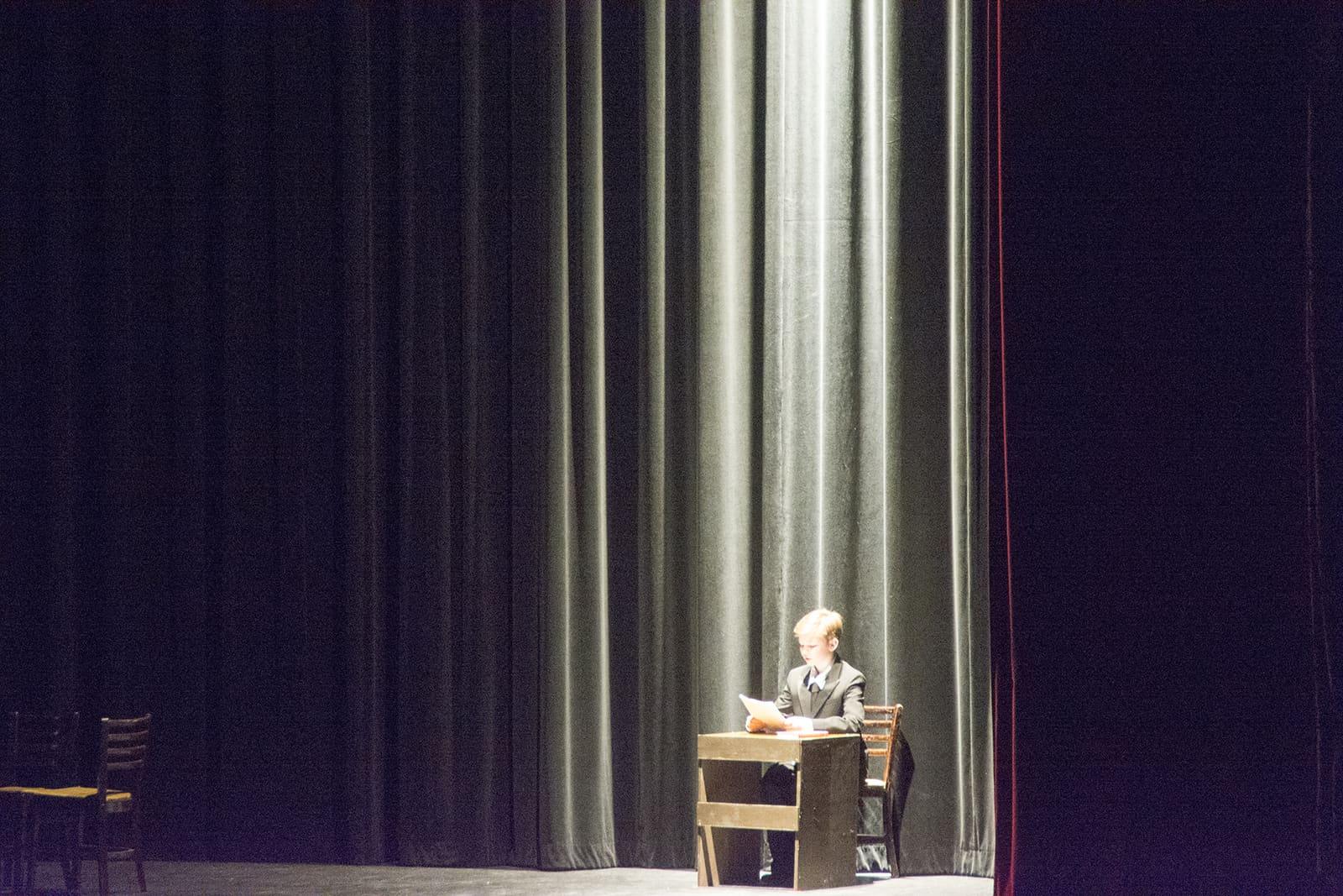 A lone speaker sits at a table on a mostly dark stage.