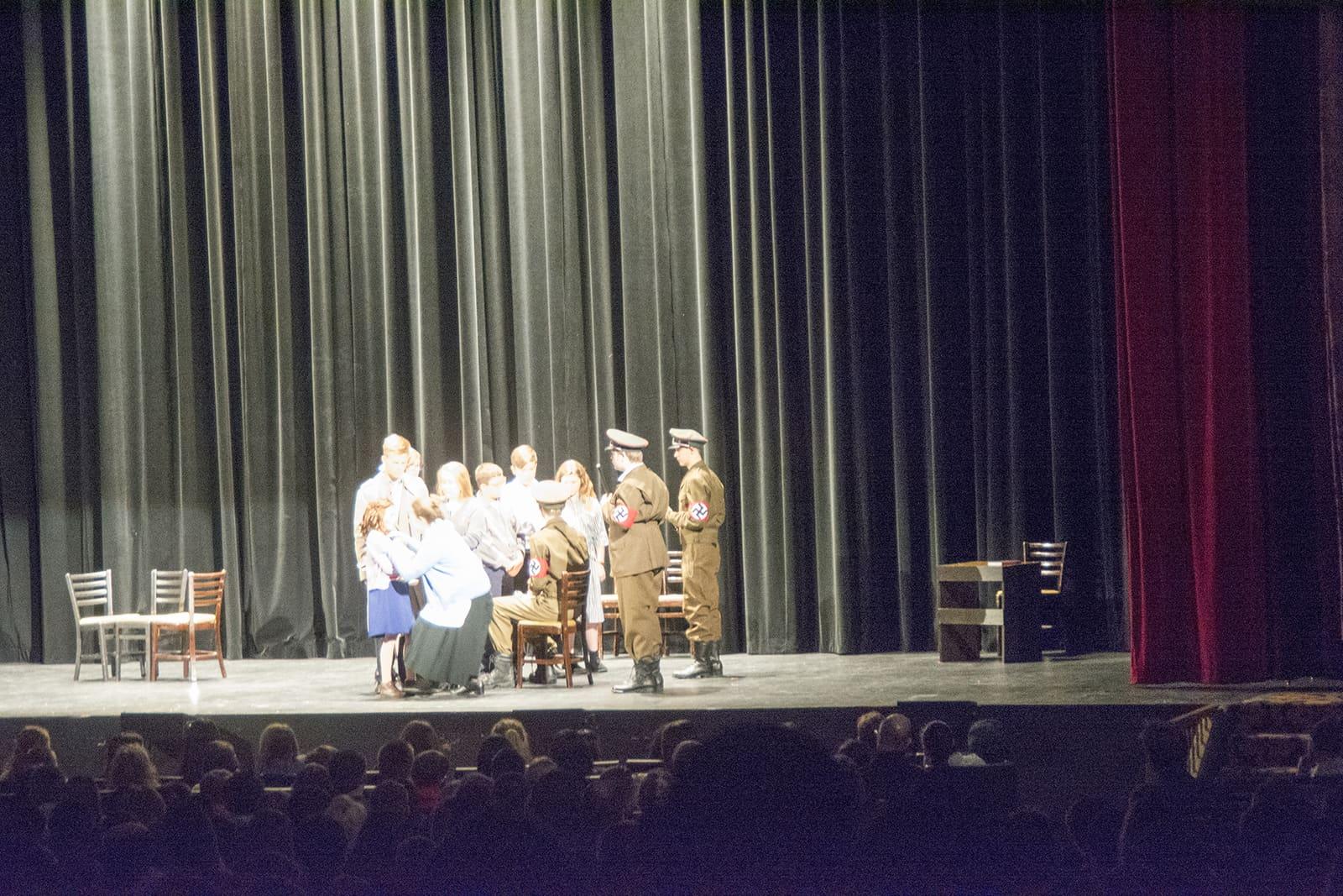 Performers gather around a table beneath tall stage curtains.