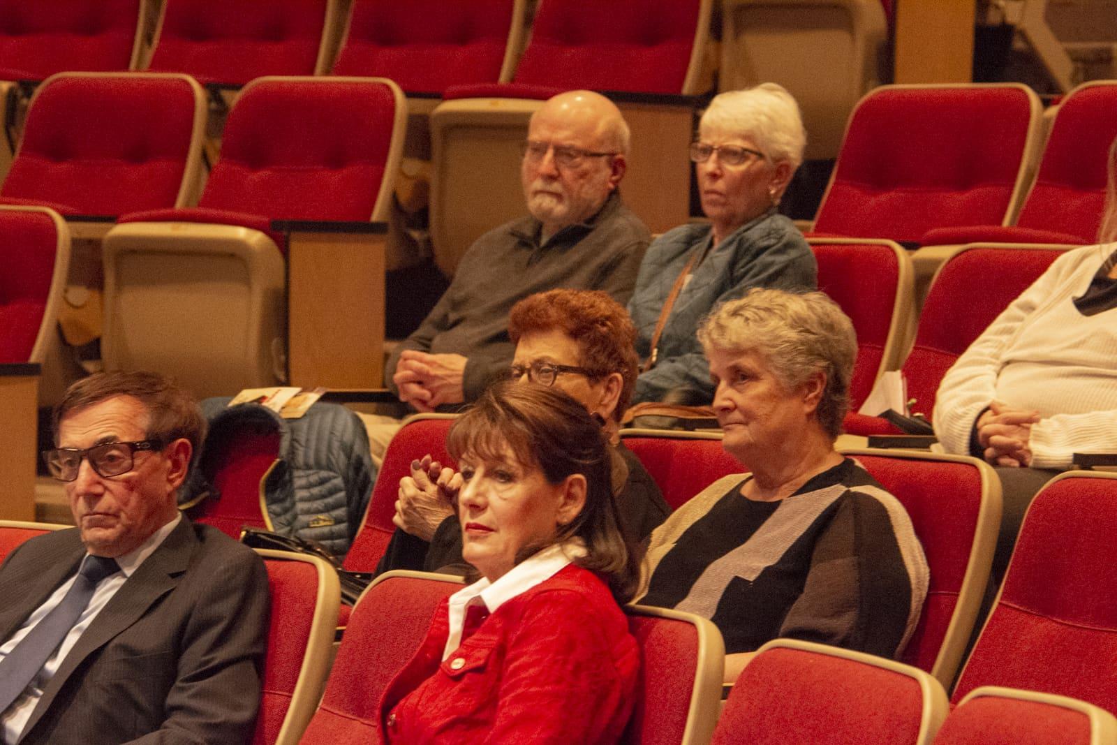 Adults converse while seated in red auditorium chairs.