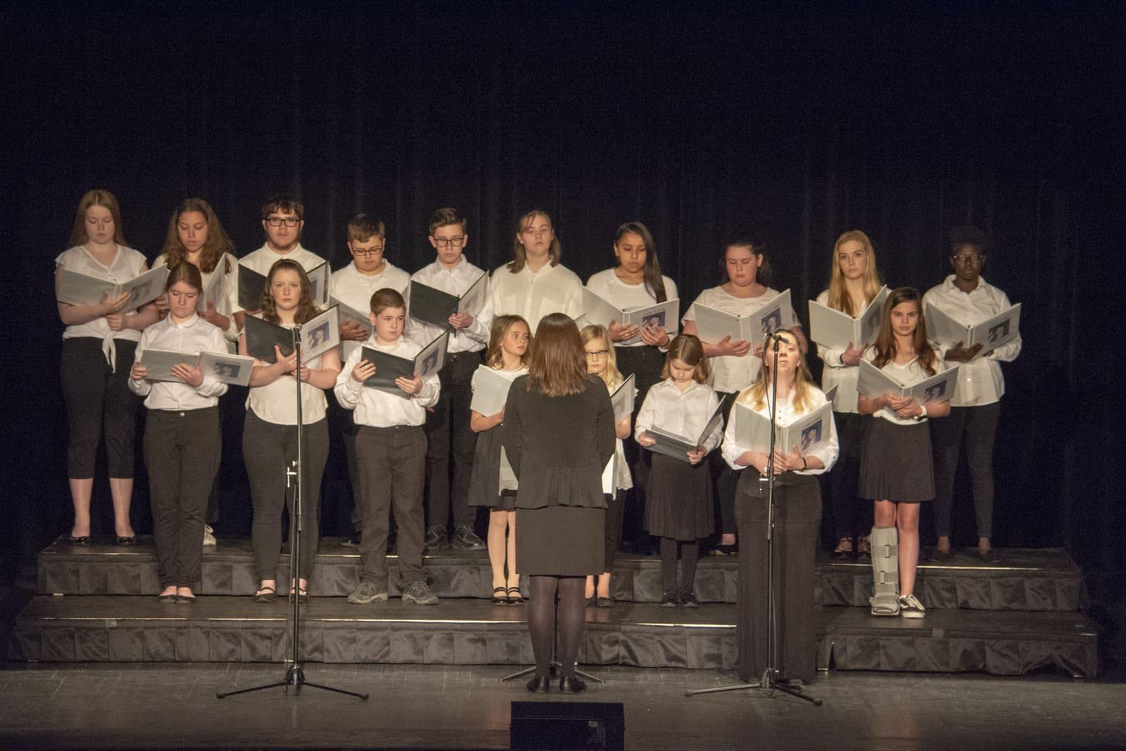 A student choir sings onstage while holding printed programs.