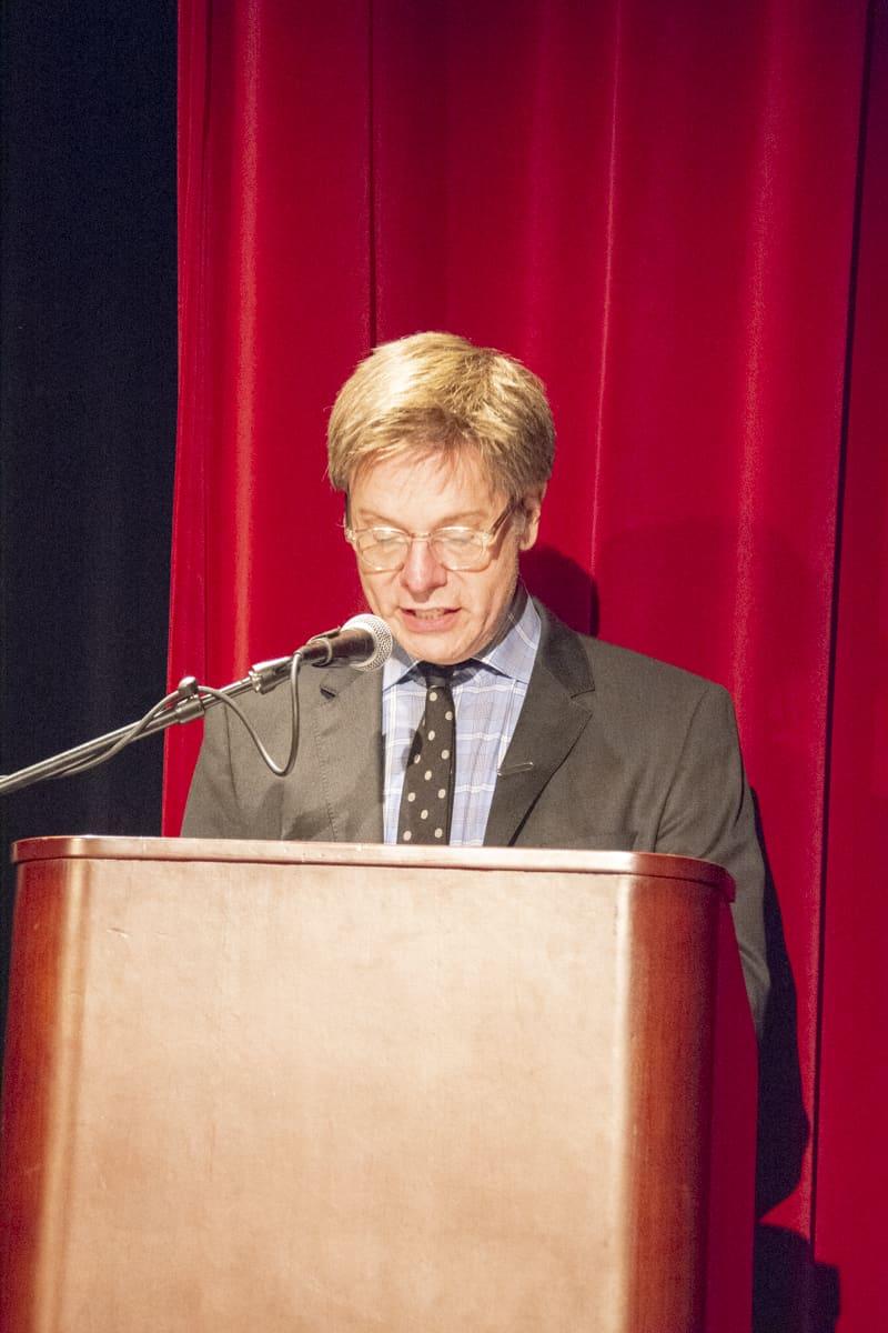 An adult reads from a lectern against a red stage curtain.