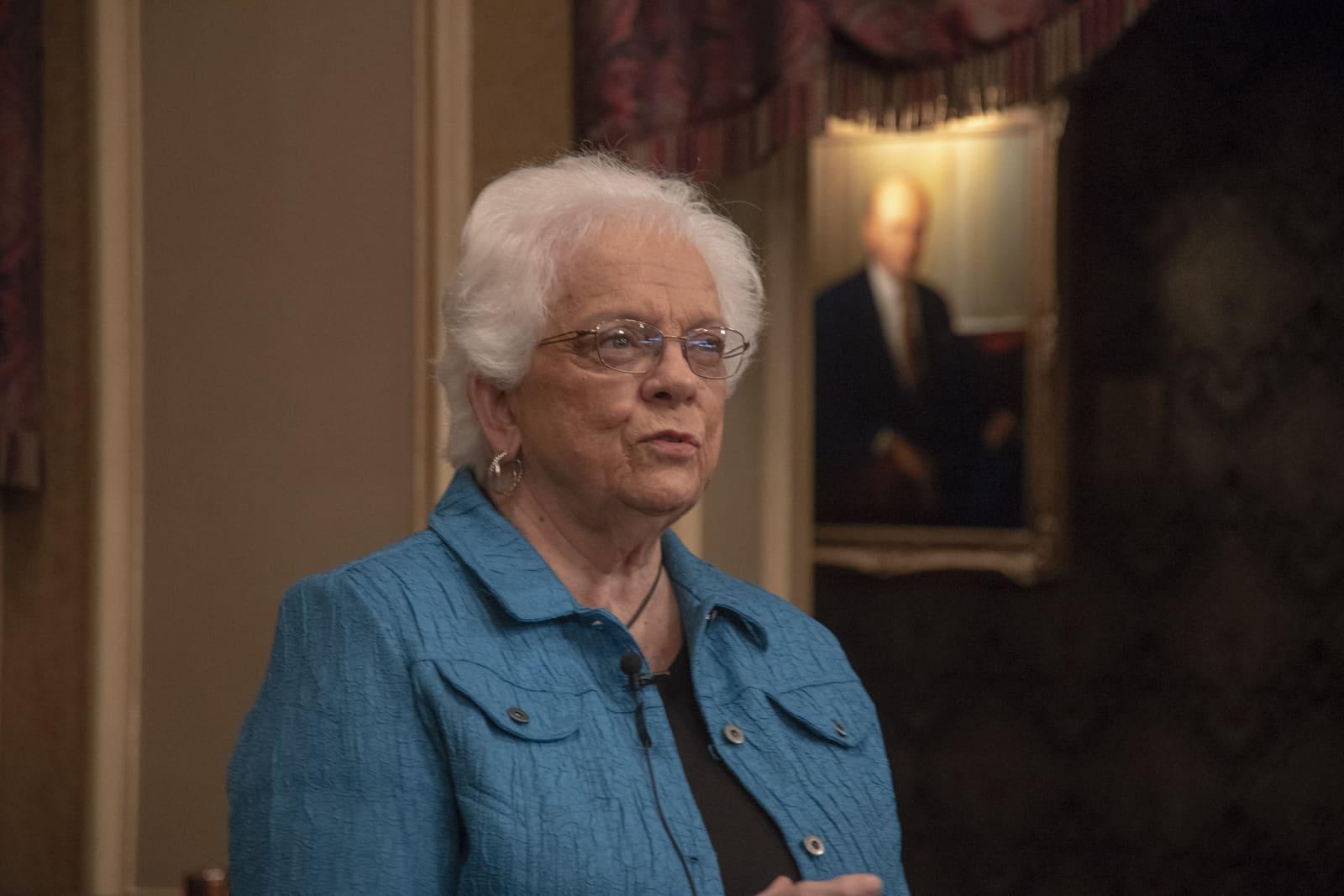 An adult sits indoors with a framed portrait behind them.