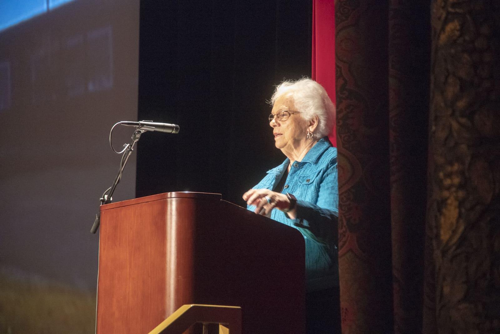 An adult speaks from a wooden lectern at the edge of a stage.