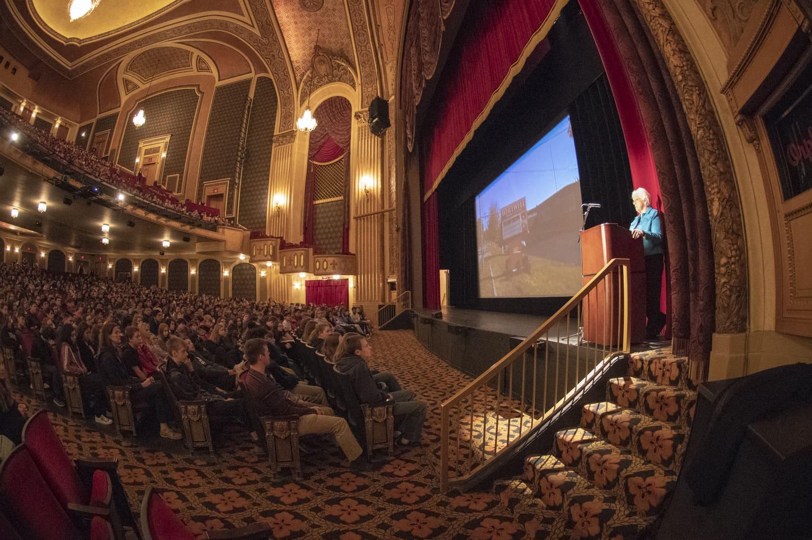 A crowded theater watches a projected speaker from beside the stage.