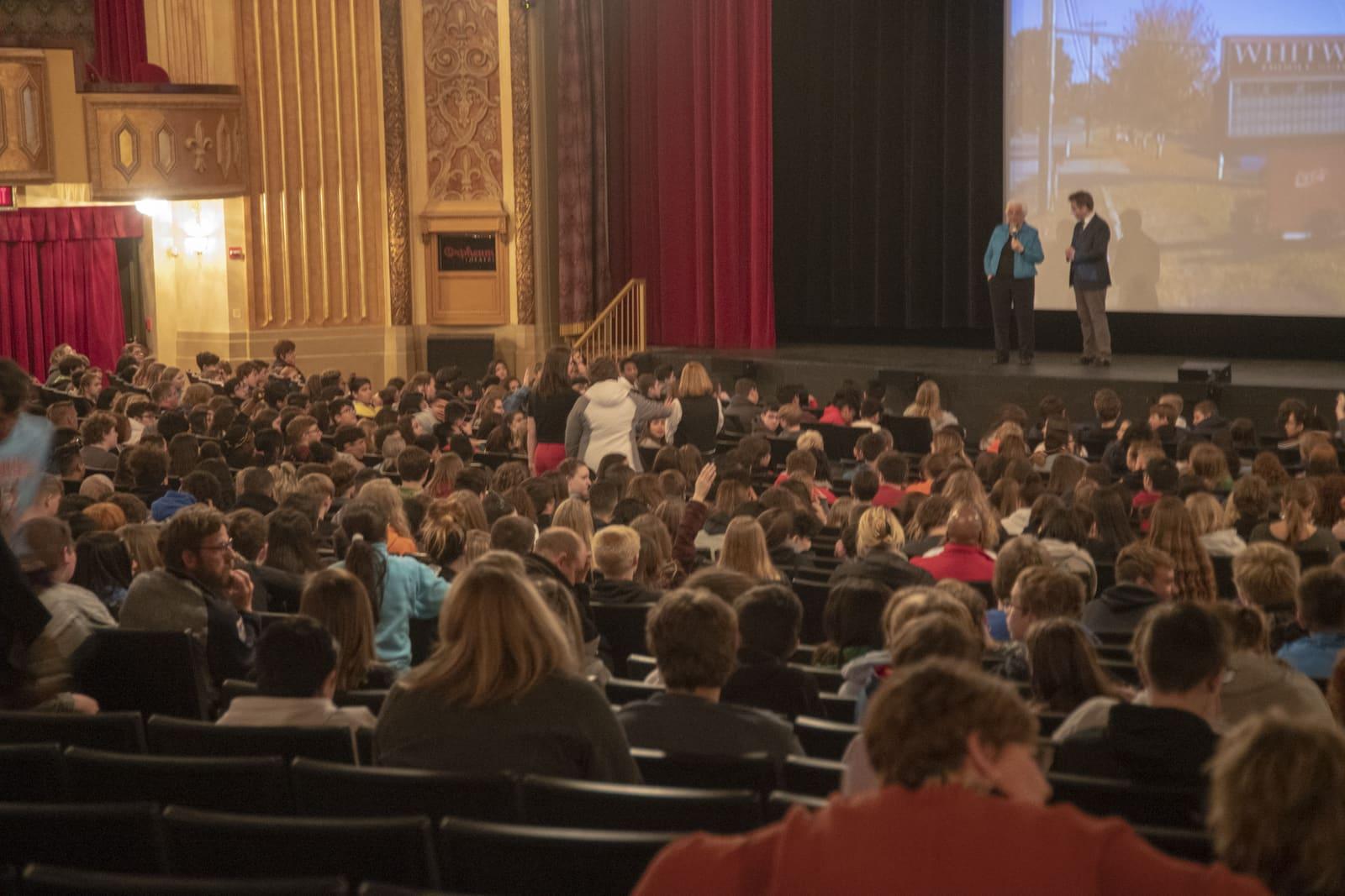 A theater audience faces two speakers onstage beside a projection.