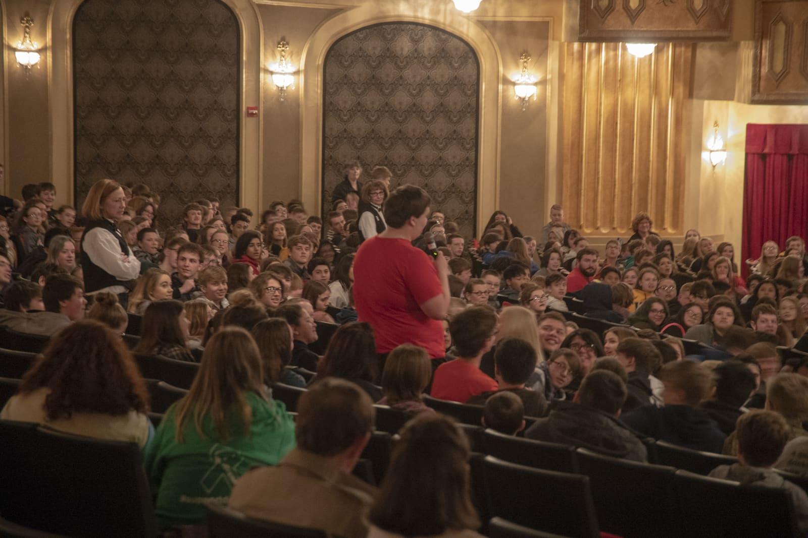 A person in red stands among a seated theater audience.