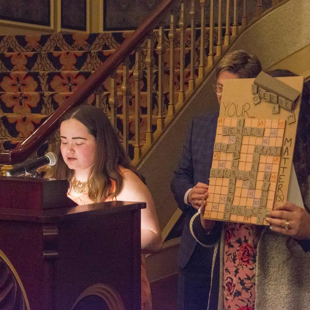 A student reads at a lectern while an adult displays grid-based artwork.