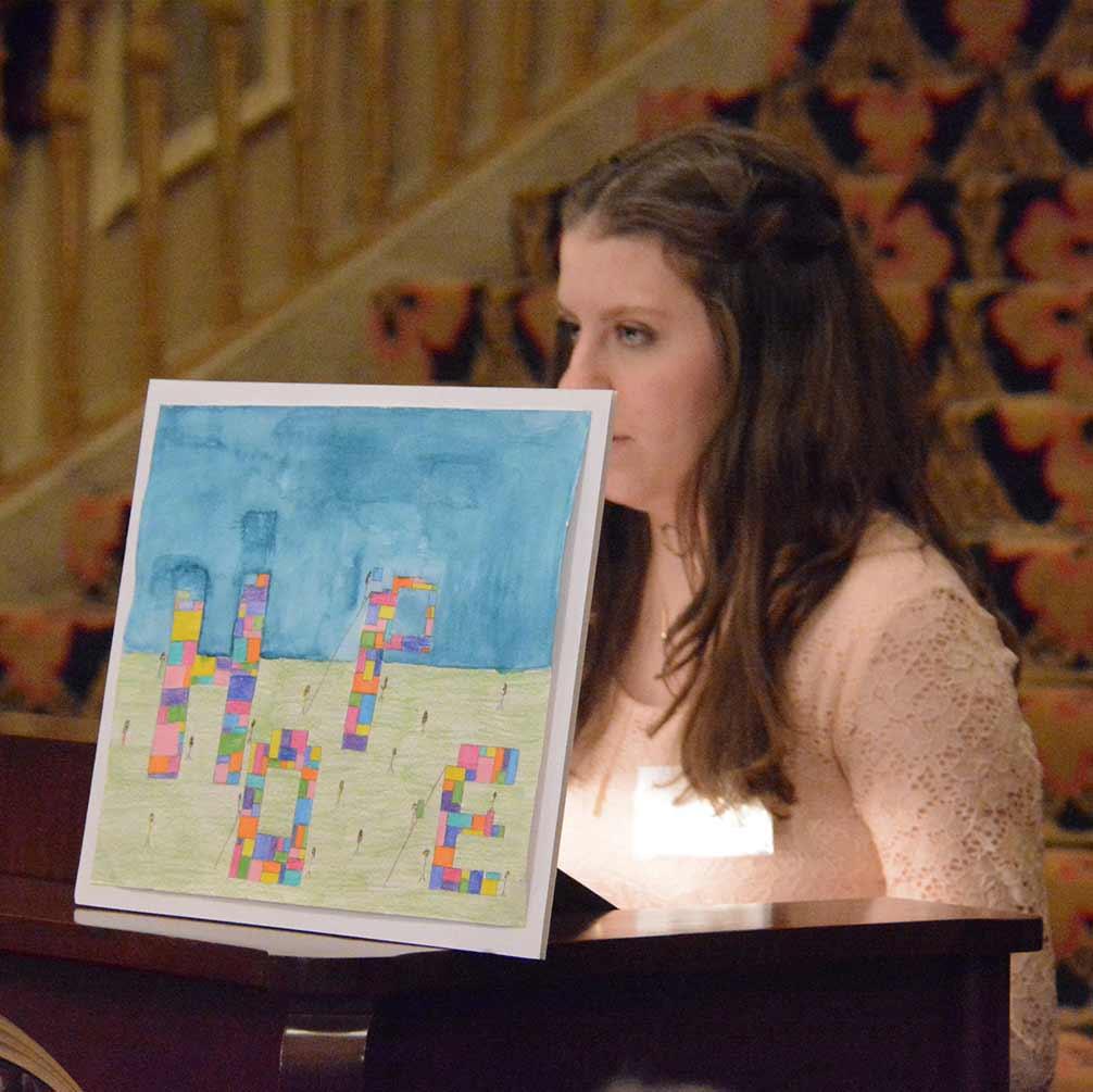 A student holds up colorful block artwork while standing at a lectern.