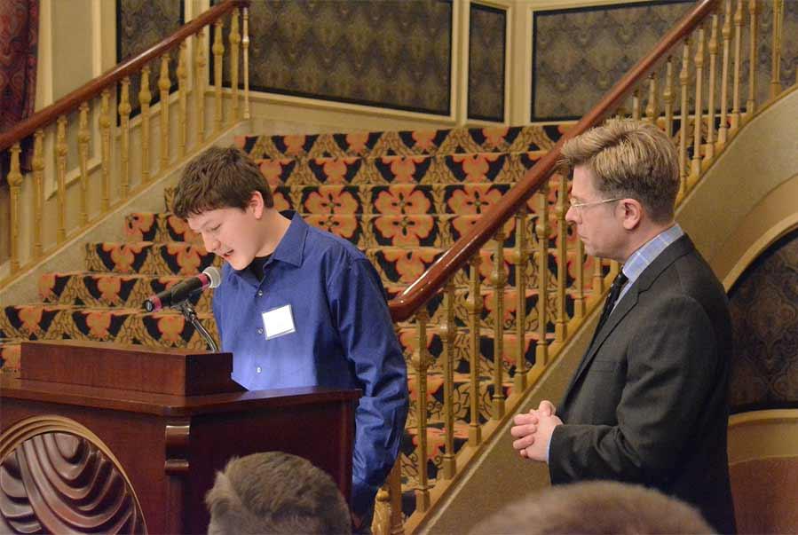 A student stands at a wooden lectern while an adult watches.