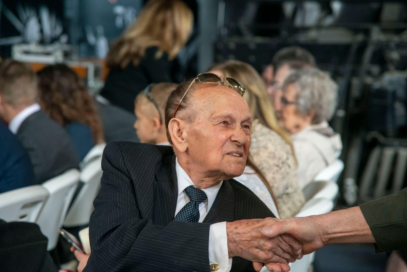 A seated adult in a suit shakes hands at an indoor gathering.