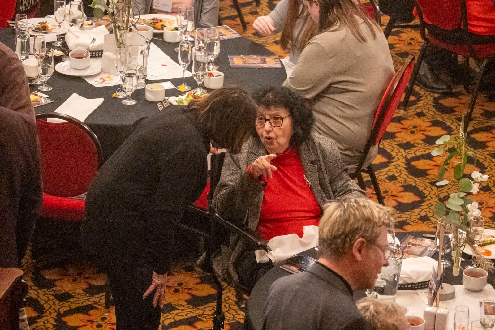 A person leans over a seated adult at a banquet table.