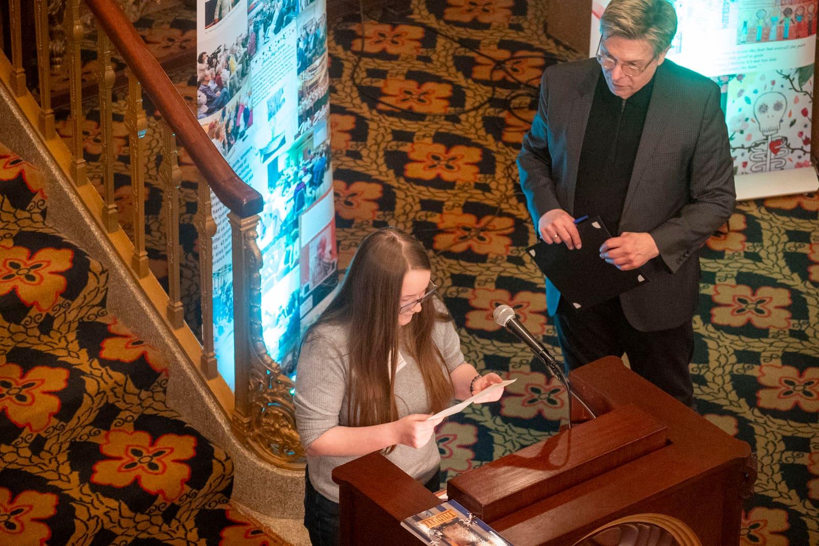 An adult reads at a lectern while an adult stands beside them.