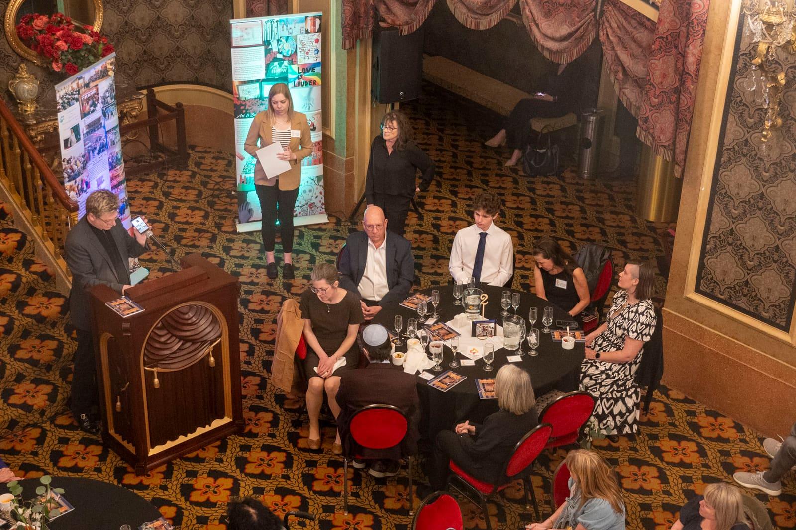A speaker stands at a lectern beside banquet tables.