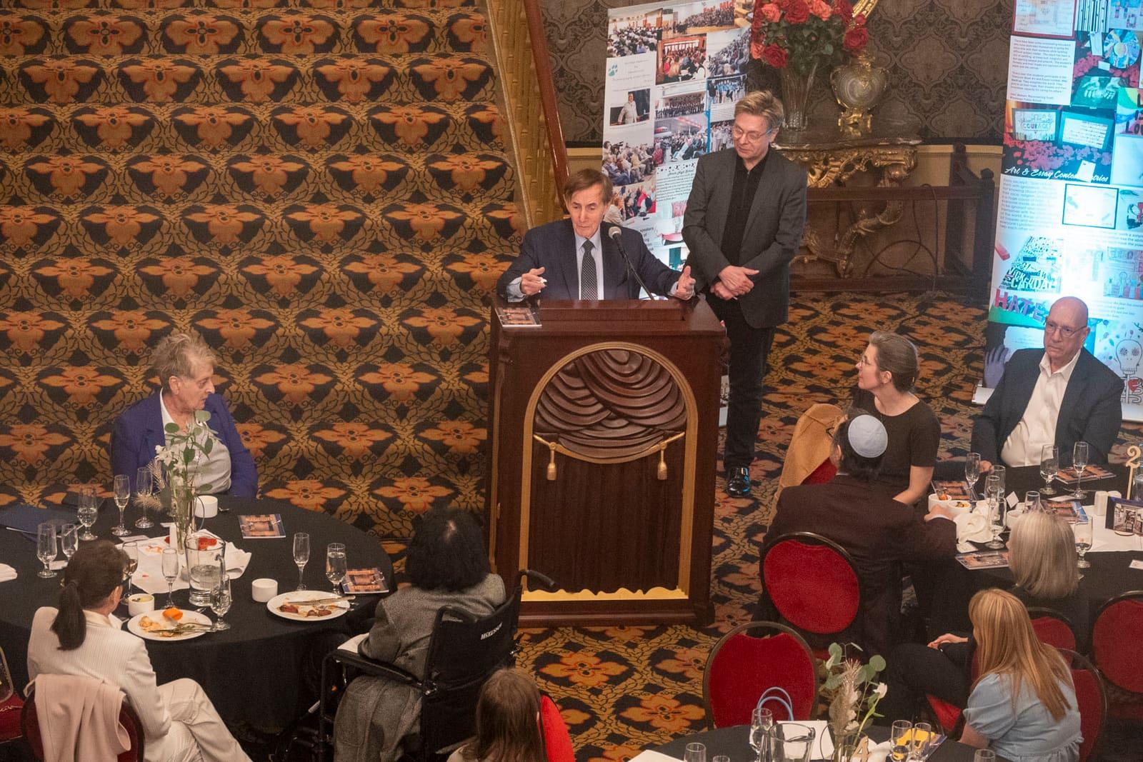 An adult speaks from a lectern while seated guests watch.