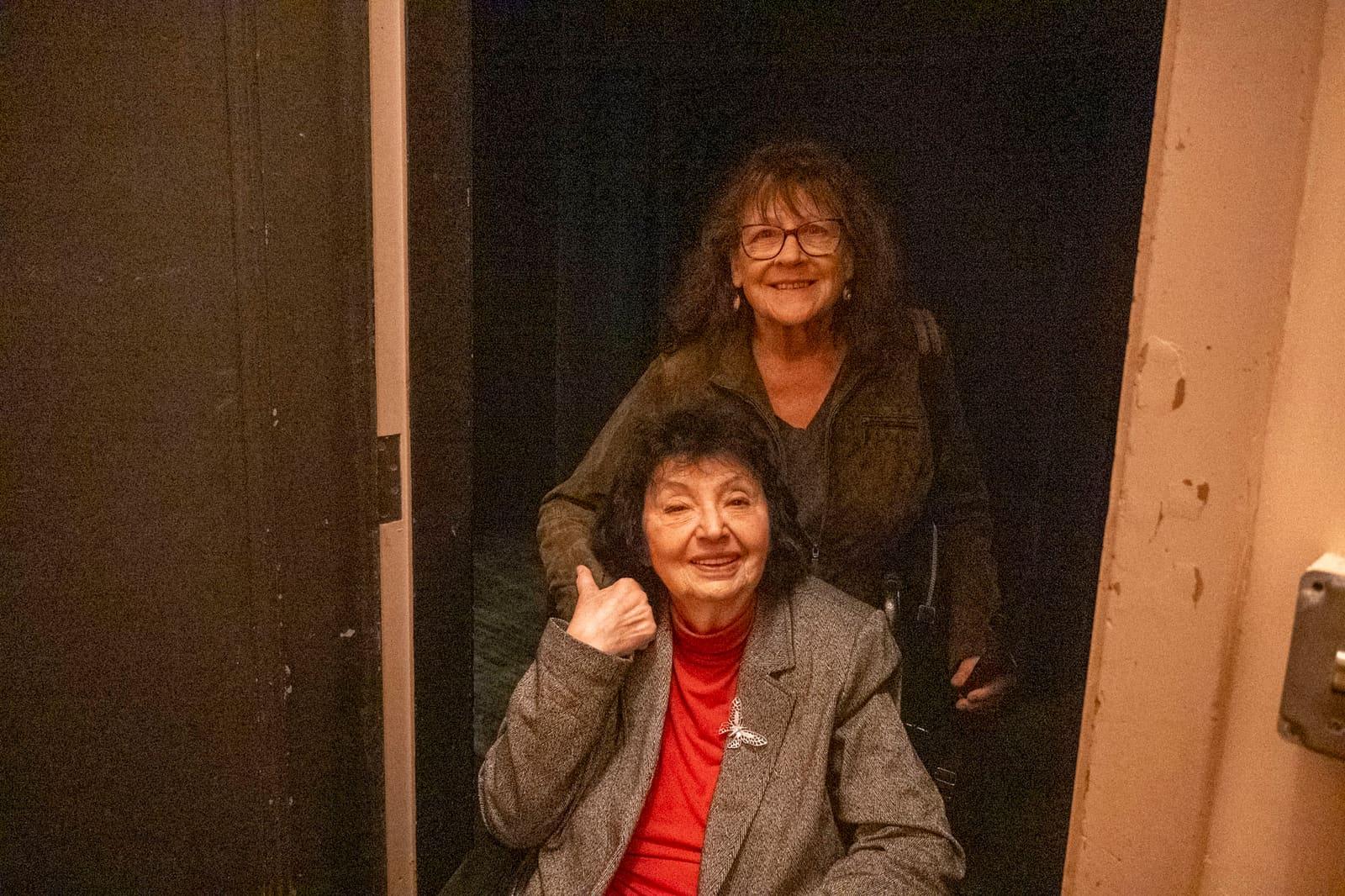 Two adults pose backstage as the seated adult gives a thumbs-up.