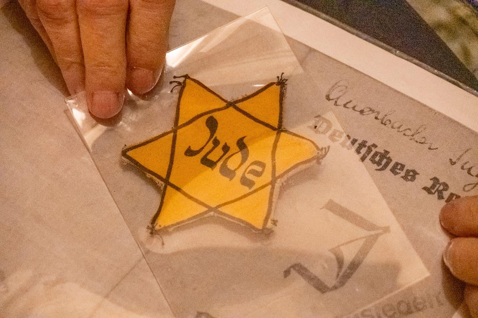 A hand holds an embroidered yellow star marked “Jude” over old documents.