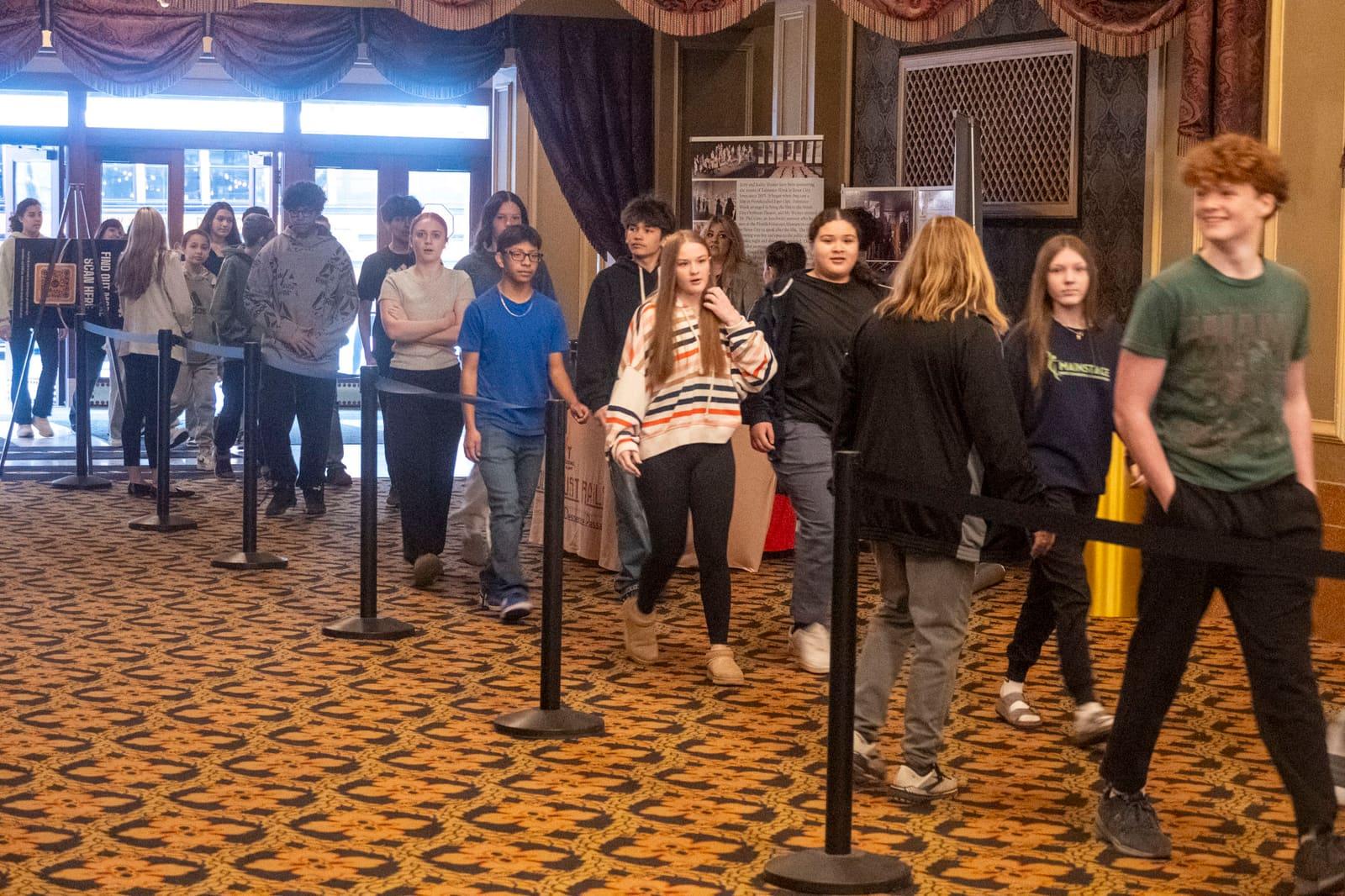 Students queue between rope barriers in a theater lobby.