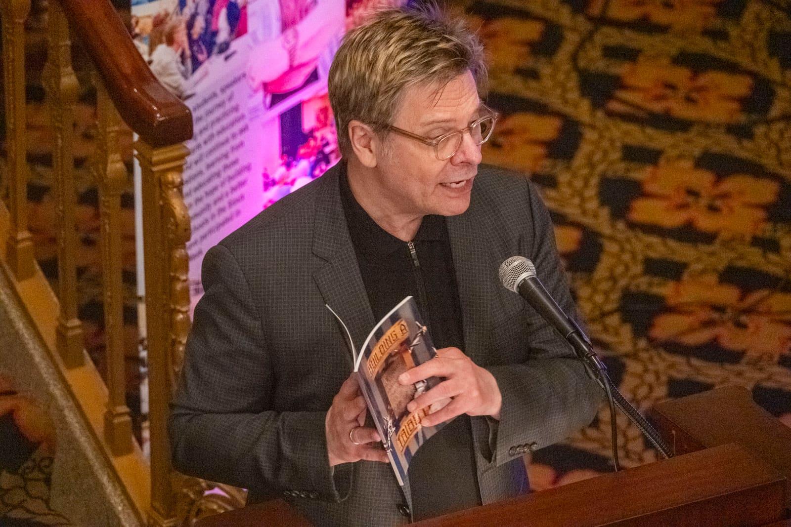An adult at a lectern holds up a printed booklet.