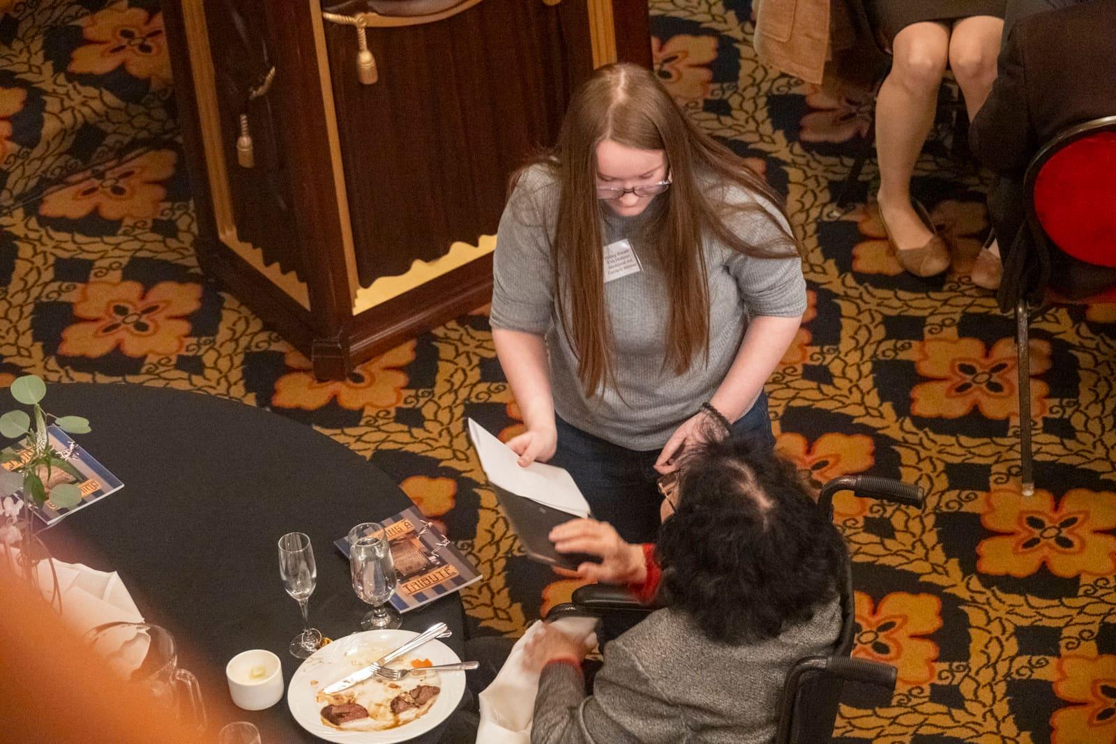 A student kneels beside a seated guest at a banquet table.