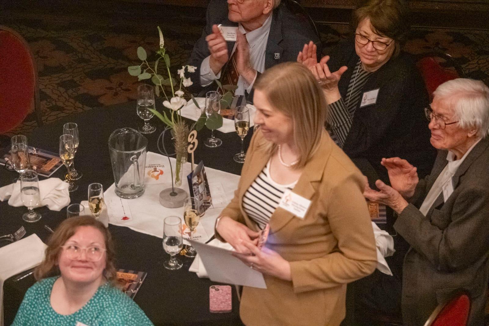 An adult stands reading a paper as nearby guests applaud.