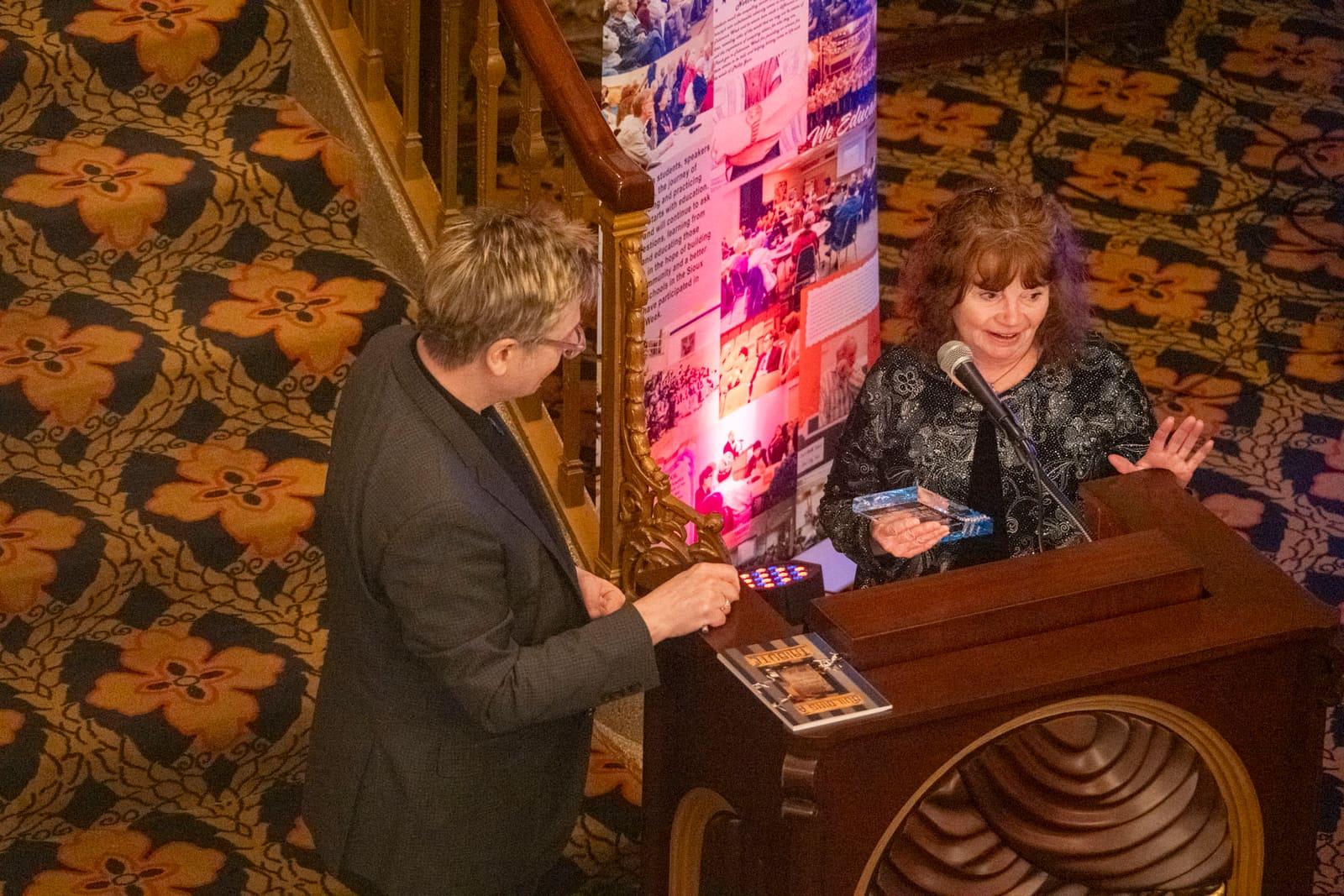 An adult presents a booklet to an adult at a lectern.