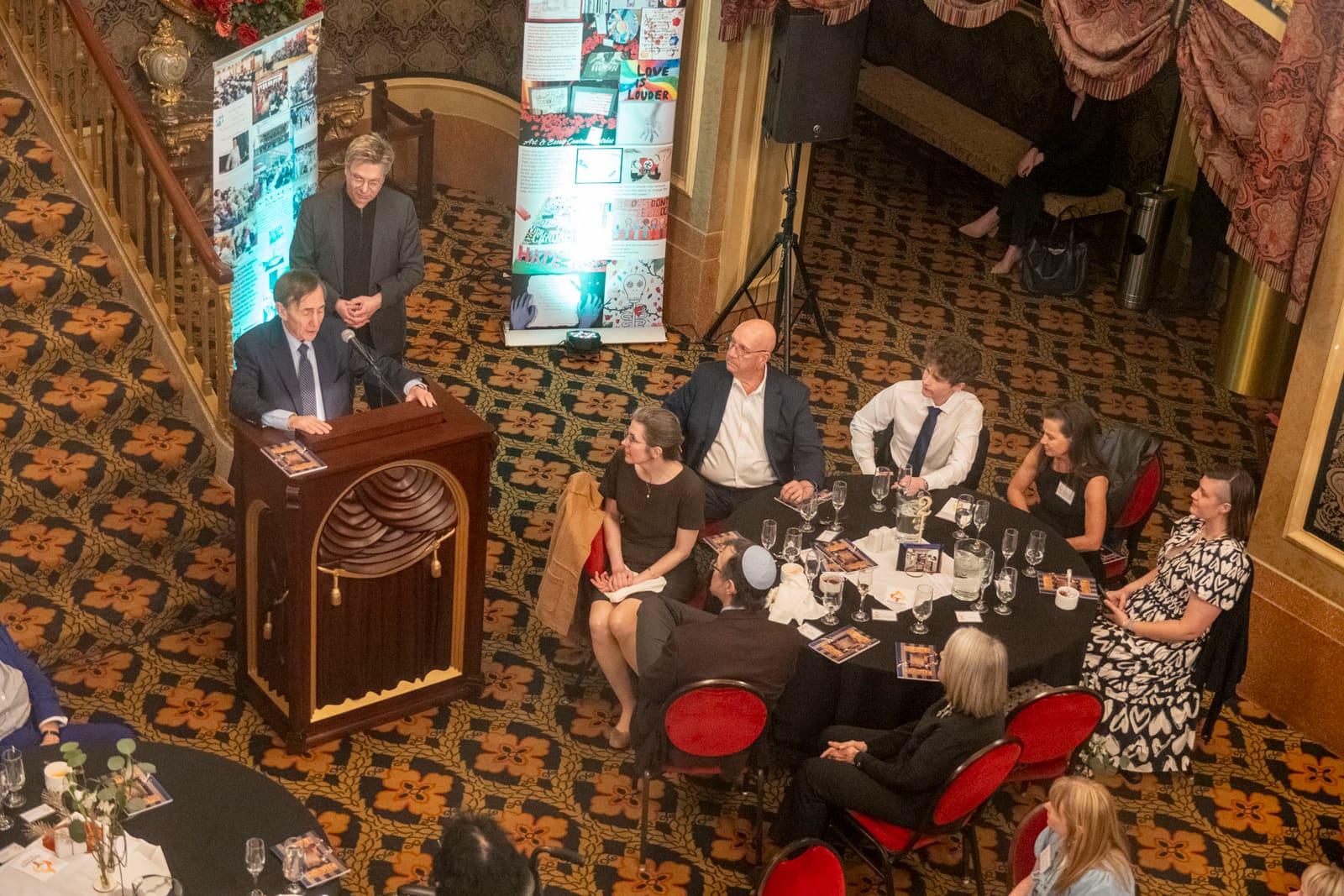 An adult speaks from a lectern beside seated banquet guests.
