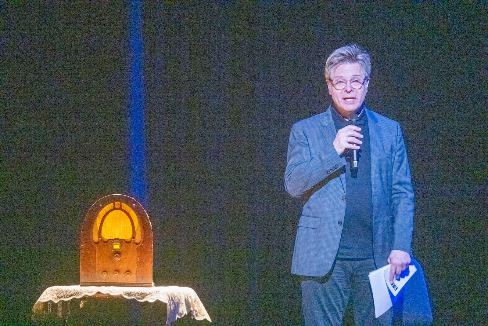 An adult speaks onstage beside a vintage wooden radio.