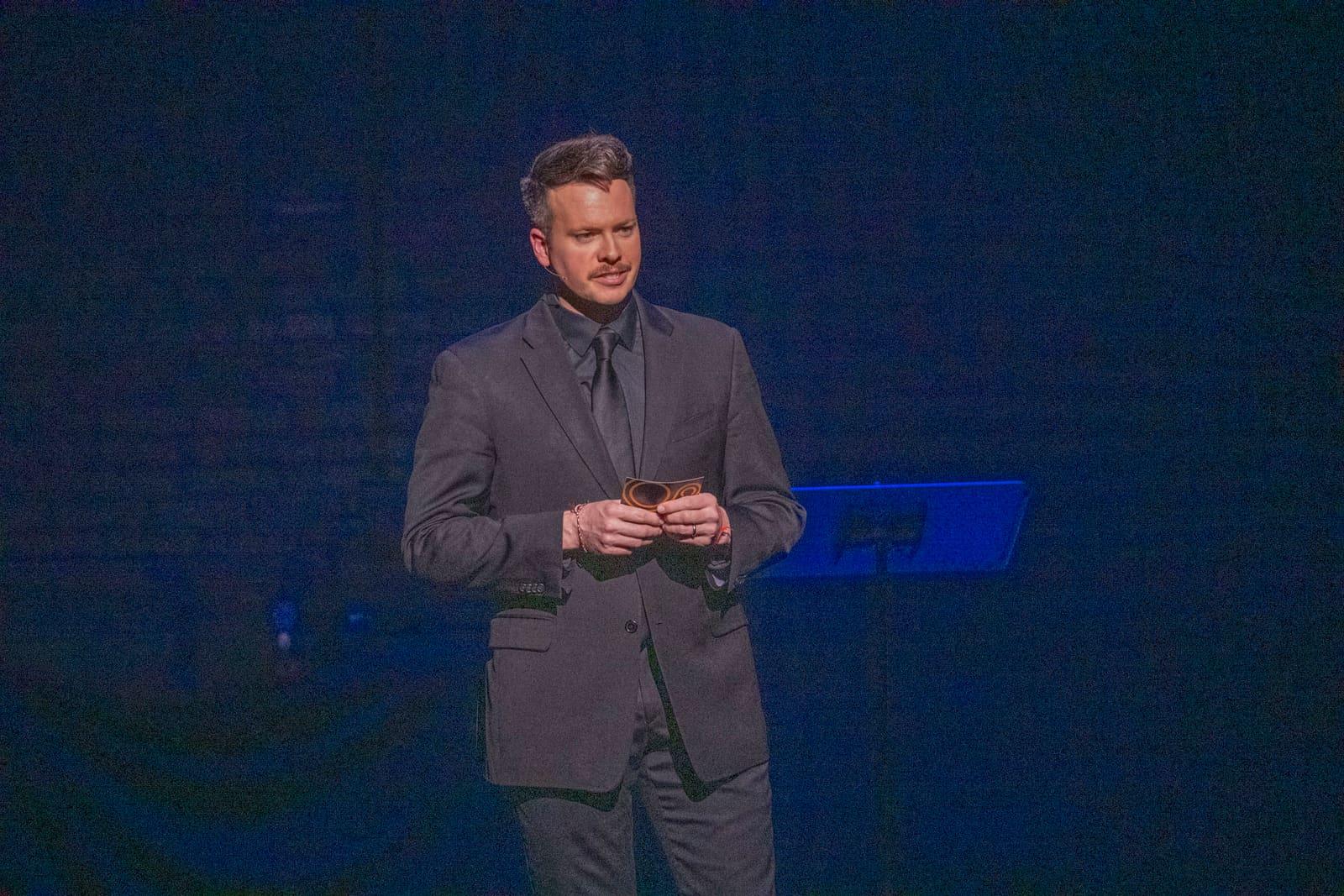 An adult stands alone on a dark stage holding note cards.