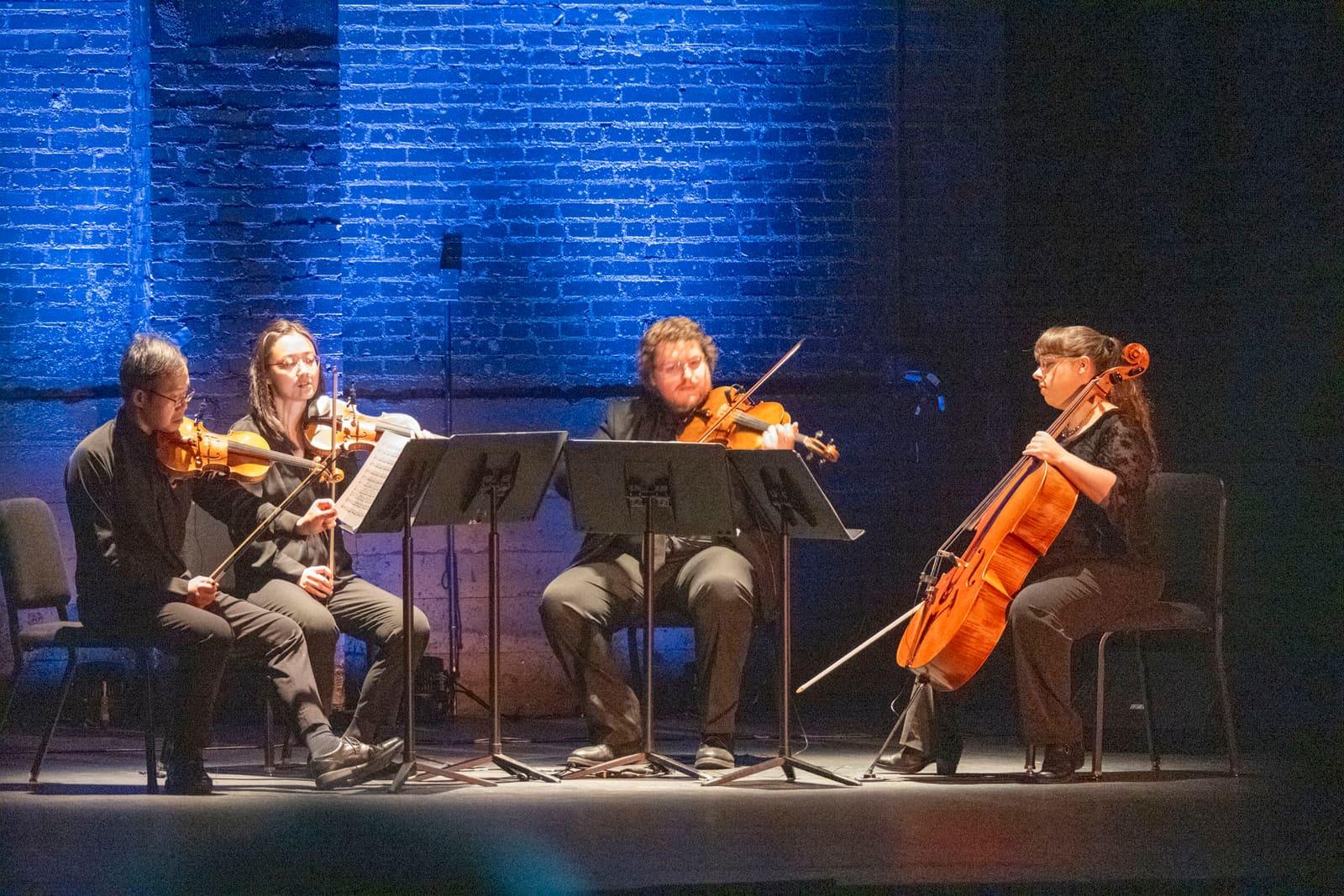 Four string musicians perform while seated onstage.