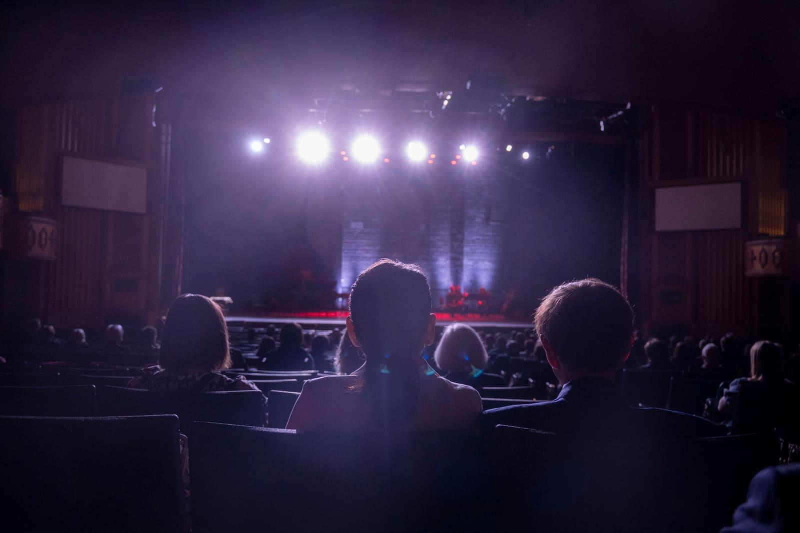 Audience silhouettes face bright lights on a dark stage.