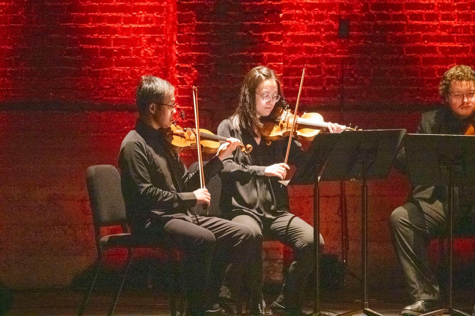 Two violinists perform against a red-lit brick wall.